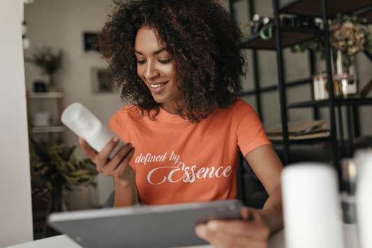 Women wearing a orange signature Defined By Essence T-Shirt with white writing, looking at a bottle